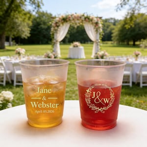 Peut inclure: Deux gobelets en plastique transparents remplis de boissons, une ambrée et une rouge, posés sur une table blanche. La boisson ambrée contient des glaçons et le texte "Jane & Webster April 05.2026". La boisson rouge a une couronne dorée avec "J&W".