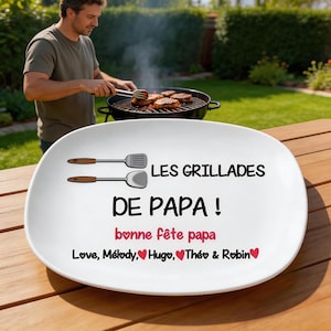 Peut inclure: Un plat de service ovale blanc orné de deux spatules en métal illustrées et du texte noir LES GRILLADES DE PAPA ! suivi du texte rose bonne fête papa et des prénoms Love, Mélody, Hugo, Théo & Robin accompagnés de cœurs rouges.