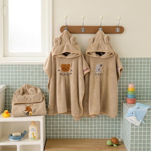 Peut inclure: Deux ponchos de bain à capuche beiges avec broderies d'ours et de lapin suspendus à une patère en bois, accompagnés d'une serviette à capuche pliée avec broderie de faon, d'un gant de toilette dauphin bleu et de jouets de bain.