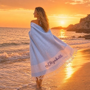 Peut inclure: Une serviette de plage texturée bleu clair et blanche avec une bande horizontale blanche près du bord inférieur. Le prénom Elizabeth est brodé dans une police cursive foncée sur la bande blanche, accompagné d'un petit cœur. La serviette a des franges.