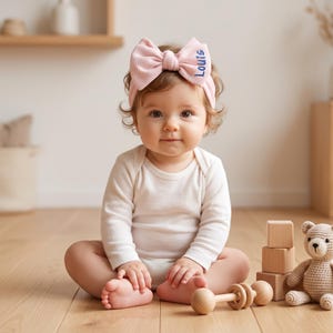 Könnte beinhalten: Ein hellrosa Stirnband mit Waffelstruktur, einer großen Schleife und dem in blauem Garn gestickten Namen Louis. Das Stirnband wird von einem Baby getragen, das einen schlichten weißen Langarmbody trägt, der weichen Stoff und ein einfaches Design zeigt.