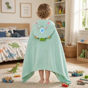 Peut inclure: Une serviette à capuche vert clair présentant un visage de monstre sur la capuche avec un œil bleu, des accents roses et des détails texturés verts. La serviette est drapée sur le dos d'un enfant, montrant un tissu doux et une forme ample.