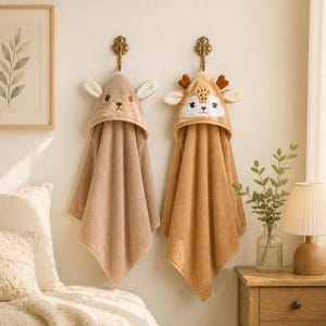 Peut inclure: Deux serviettes à capuche suspendues à des crochets muraux. La serviette de gauche est marron clair avec des oreilles et un visage brodés. La serviette de droite est de couleur fauve avec un visage de cerf détaillé, des bois et des oreilles.