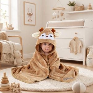 Peut inclure: Une serviette à capuche douce de couleur beige, ornée d'un motif de visage de cerf avec des oreilles et des bois marron. Le tissu en peluche porte le nom Ahern brodé en écriture bleue dans le coin inférieur, enveloppant confortablement l'enfant.