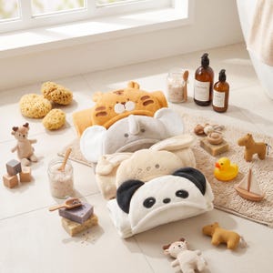 Peut inclure: Une pile de quatre serviettes à capuche sur le thème des animaux, représentant un tigre jaune, un éléphant gris, un lapin crème et un panda blanc et noir. Les serviettes sont disposées sur un tapis de bain beige avec des accessoires et jouets.