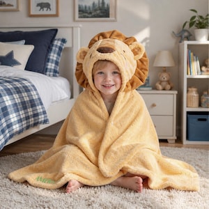 Peut inclure: Une serviette à capuche jaune douce avec un visage de lion en peluche, un nez marron et une crinière texturée. Le tissu a un aspect polaire duveteux et le mot AMERIS est brodé en fil vert dans le coin inférieur de la couverture.