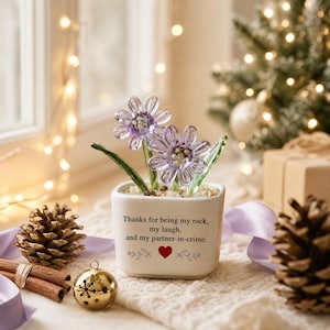 Peut inclure: Un pot en céramique blanc de forme carrée contenant deux fleurs en verre violet avec des tiges vertes. Le pot porte l'inscription noire : Thanks for being my rock, my laugh, and my partner-in-crime, avec un petit cœur rouge et des feuilles.