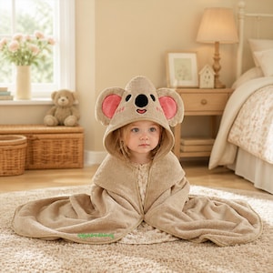 Peut inclure: Une serviette à capuche beige arborant un motif de visage de koala avec de grandes oreilles arrondies, des accents roses à l'intérieur des oreilles, un nez noir et des yeux brodés. Le tissu doux et pelucheux enveloppe les épaules.