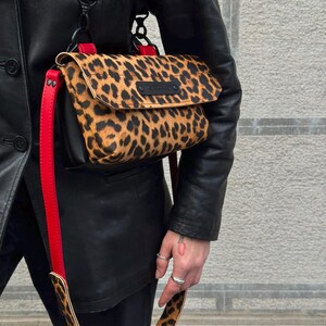 Pode incluir: Uma bolsa tiracolo com aba de estampa de leopardo, painéis laterais pretos e uma alça ajustável em vermelho vibrante. A bolsa inclui ferragens pretas, uma etiqueta de marca em couro preto na aba e uma alça de ombro a condizer com estampa.