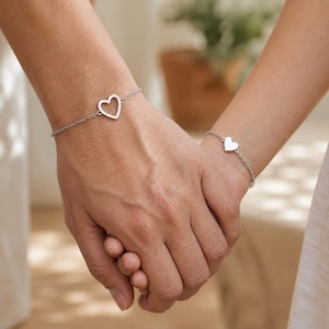 Könnte beinhalten: Zwei silberfarbene Kettenarmbänder, die jeweils mit einem herzförmigen Anhänger versehen sind. Ein Armband zeigt eine offene Herzform, während das andere Armband einen massiven, ausgefüllten Herzanhänger aufweist. Beide Stücke bestehen aus feinen Metallgliedern.