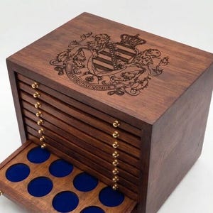 Pode incluir: Um gabinete de madeira castanho-escuro para moedas com oito gavetas horizontais e pequenos puxadores dourados. A superfície superior apresenta um brasão heráldico gravado a laser com uma coroa e o texto em latim PROVIDENTIAE MEMOR. A gaveta inferior está aberta, revelando um interior forrado a veludo azul com cavidades circulares para moedas.