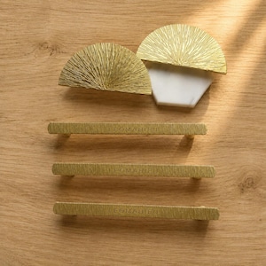 May include: Three gold-toned metal drawer pulls with a textured finish and the engraved text LEOMAHIEU, alongside two semi-circular gold-toned metal cabinet knobs, one of which is mounted on a white hexagonal marble base with a textured surface.