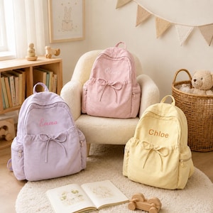 May include: Three fabric backpacks in light purple, soft pink, and pale yellow. Each backpack features a front pocket with a drawstring bow, side pockets with zippers, and a name embroidered on the front: Emma, Olivia, and Chloe respectively.