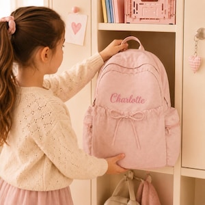 May include: A light pink corduroy backpack featuring the name Charlotte embroidered in a matching pink script font on the front pocket. The backpack includes a top handle, two side pockets with zips, and a decorative bow detail on the front.