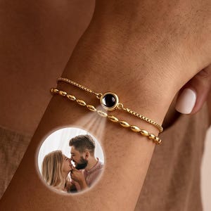 Puede incluir: Una pulsera dorada de doble cadena con un dije circular central que proyecta una pequeña imagen fotográfica en blanco y negro de una pareja abrazándose. La pulsera consta de una cadena fina y otra cadena con pequeñas cuentas ovaladas.