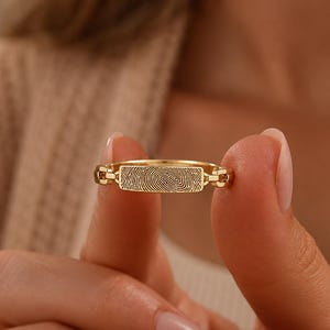 Op de afbeelding: Een goudkleurige ring met een rechthoekige middenplaat gegraveerd met een gedetailleerd vingerafdrukpatroon. De band bestaat uit kleine, onderling verbonden schakels aan weerszijden van de centrale plaat voor een unieke textuur.