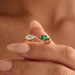 Op de afbeelding: Een gouden ring met twee marquise-geslepen stenen, één heldere en één smaragdgroene, gezet in een bypass-stijl. De ringband is dun en van gepolijst goud, wat een strak ontwerp laat zien dat de contrasterende kleuren van de stenen benadrukt.
