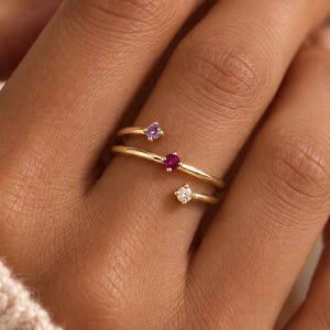 May include: A gold bypass ring featuring three small round-cut gemstones. The ring has an open design with a purple stone at one end, a red stone in the center, and a clear white stone at the other end, all set on a thin polished gold band.