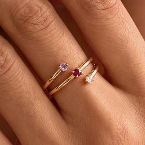 Puede incluir: Un anillo de oro tipo bypass con tres pequeñas piedras preciosas de corte redondo en diferentes colores. El anillo tiene una banda fina de oro pulido que se envuelve alrededor del dedo, con una piedra morada, una roja y una blanca en los extremos.