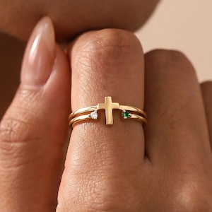 May include: A gold ring featuring a central cross design flanked by a small clear gemstone on one side and a small green gemstone on the other. The band is thin and polished, showcasing a minimalist aesthetic with a delicate metal construction.