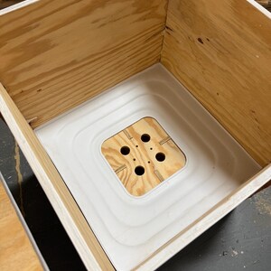 Pu&ograve; includere: Una scatola di legno con interno bianco e base quadrata in legno con quattro fori. La scatola &egrave; costruita in compensato di colore chiaro e ha un aspetto pulito e non rifinito. L'interno bianco ha un design a gradini.