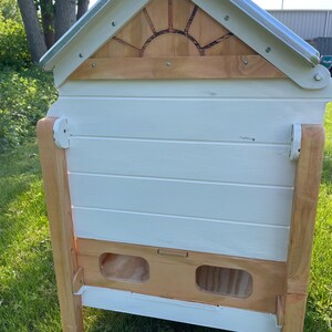 Pu&ograve; includere: Un'arnia in legno verniciata di bianco con cornice e dettagli in legno marrone chiaro. Il tetto &egrave; argentato e la parte anteriore presenta due aperture. La parte superiore dell'arnia presenta un disegno a raggiera. L'arnia &egrave; all'aperto su una superficie erbosa.