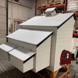 Pu&ograve; includere: Un pollaio in legno bianco con tetto in metallo ondulato e finiture nere. Il pollaio presenta una piccola porta rossa, due nidi e un camino di ventilazione. Il pollaio &egrave; costruito su un telaio di legno.