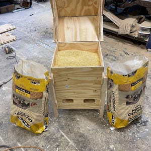 Pu&ograve; includere: Un mangiatoia per polli in legno riempita di mais spezzato, affiancata da due sacchi di mais spezzato. La mangiatoia &egrave; costruita in compensato di colore chiaro e ha un coperchio incernierato. I sacchi sono gialli e presentano una grafica di anatre.