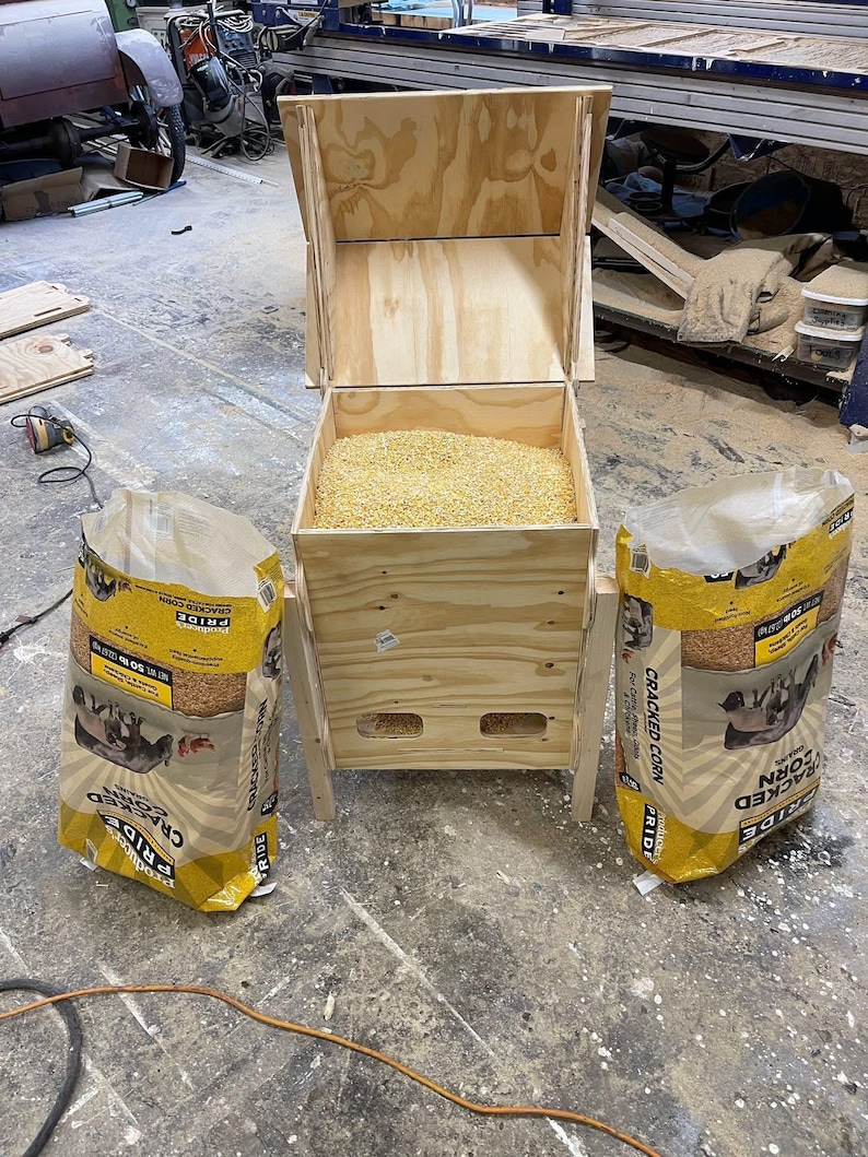Pu&ograve; includere: Un mangiatoia per polli in legno riempita di mais spezzato, affiancata da due sacchi di mais spezzato. La mangiatoia &egrave; costruita in compensato di colore chiaro e ha un coperchio incernierato. I sacchi sono gialli e presentano una grafica di anatre.