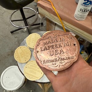 Pu&ograve; includere: Una placca decorativa color rame con la scritta "MADE IN LAPEER MI USA" e "KritterKribs.com". Sono visibili anche diverse placche beige pi&ugrave; piccole simili. Una bomboletta di vernice trasparente lucida &egrave; sullo sfondo.