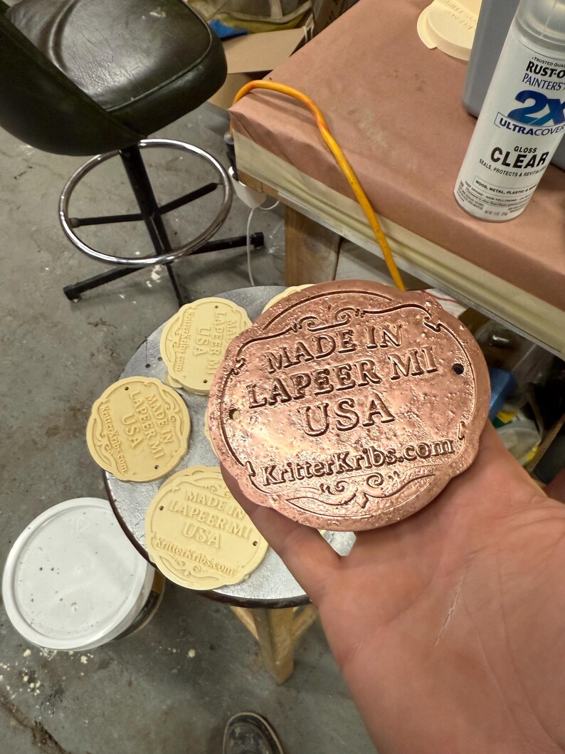 Pu&ograve; includere: Una placca decorativa color rame con la scritta "MADE IN LAPEER MI USA" e "KritterKribs.com". Sono visibili anche diverse placche beige pi&ugrave; piccole simili. Una bomboletta di vernice trasparente lucida &egrave; sullo sfondo.