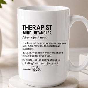 May include: A white ceramic mug featuring black printed text that defines a therapist as a mind untangler. The text includes a phonetic pronunciation, a noun label, and three numbered descriptions of professional duties, ending with the name Tyler.