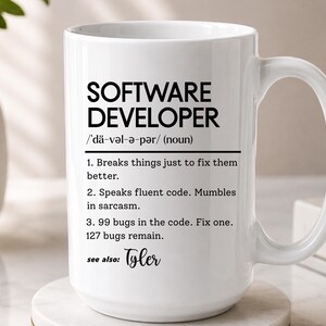 Pode incluir: Uma caneca de cerâmica branca com texto impresso em preto que define um desenvolvedor de software. O texto inclui uma pronúncia fonética, um rótulo de substantivo e três pontos humorísticos numerados sobre codificação, terminando com Tyler.