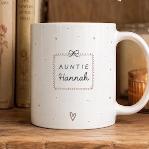 Può includere: Una tazza in ceramica bianca con una cornice rettangolare centrale sormontata da un piccolo fiocco. All'interno della cornice, il testo recita AUNTIE Hannah in caratteri neri. La tazza è decorata con piccoli cuori e puntini sparsi.