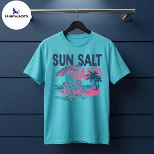 Puede incluir: Una camiseta de manga corta color turquesa con un estampado gráfico de dos sillas de playa bajo una sombrilla a rayas sobre arena rosa. El texto dice SUN SALT & SAND en letras azul oscuro y rosa, con palmeras y olas de mar de fondo.