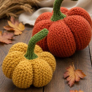 May include: Two hand-knitted pumpkins, one in a vibrant orange and the other in a golden yellow, each featuring a textured, ribbed surface and a prominent, upright green stem. The pumpkins are crafted with a visible knit stitch pattern.
