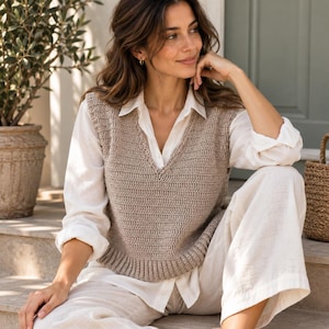 May include: A beige sleeveless crochet sweater vest with a V-neckline and ribbed hem, worn over a white long-sleeved button-down shirt and matching white wide-leg trousers. The vest features a consistent horizontal stitch pattern throughout.