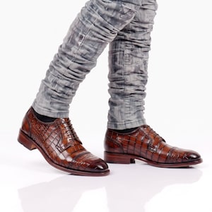 May include: A pair of brown leather oxford shoes featuring a textured crocodile skin pattern, dark brown laces, and a cap toe design. The shoes are worn with grey, vertically textured trousers that feature a subtle, mottled wash pattern.