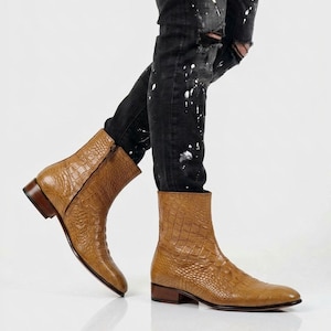Può includere: Stivaletti color cuoio con una texture a motivo pelle di coccodrillo, chiusura con zip laterale e tacco basso. Gli stivali sono indossati con jeans neri consumati che presentano schizzi di vernice bianca su tutto il tessuto.