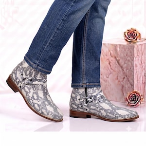 Puede incluir: Botines con estampado de piel de serpiente en blanco y negro, correa lateral con detalle de hebilla metálica, tacón bajo de madera y cierre de cremallera lateral, combinados con pantalones vaqueros azules.