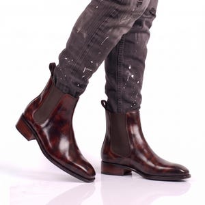 May include: A pair of dark brown, burnished leather Chelsea boots featuring a glossy, marbled finish, dark brown elastic side panels, and a pull tab at the heel. The boots have a low, stacked wooden heel and a smooth, rounded toe design.