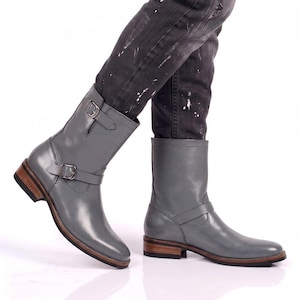 May include: A pair of mid-calf grey leather boots featuring two adjustable straps with silver-toned metal buckles, a rounded toe, and a stacked wooden heel with a black rubber sole. The boots have a smooth finish and a structured shaft design.