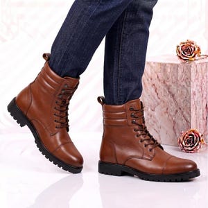 May include: A pair of brown leather lace-up boots featuring a cap toe design, a padded collar, and a thick black lug sole. The boots are worn with dark blue denim jeans, showcasing a high-top silhouette with metal eyelets and brown laces.