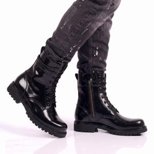 May include: A pair of black patent leather combat boots featuring a high-shine finish, black laces, a side zip closure, and a thick, lugged rubber sole. The boots have a rounded toe and a pull tab at the heel, providing a sturdy and functional design.