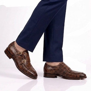 May include: A pair of brown snakeskin-patterned leather double monk strap shoes with gold-toned buckles, worn with dark navy blue trousers. The shoes feature a textured finish, a low heel, and a pointed toe design with visible stitching details.