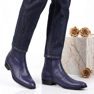 May include: A pair of blue leather ankle boots with a snakeskin texture. The boots have a side zipper and a low heel. The person is wearing dark blue jeans. Gold roses are on a marble block.