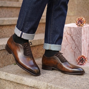 Op de afbeelding: Een paar bruine leren oxford-schoenen met donkerbruine suède vetersluiting, brogue-details op de neus en een lage hak. De schoenen worden gedragen met een donkerblauwe spijkerbroek met omgeslagen pijpen en zwarte sokken.