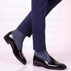 Pode incluir: Um par de sapatos de verniz preto com uma secção de tornozelo em camurça azul com quatro pequenos botões pretos na lateral. Os sapatos têm biqueira pontiaguda e salto baixo, combinados com calças azul-escuras até ao tornozelo.