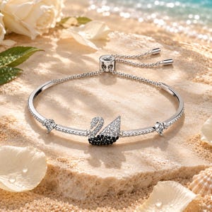 May include: A silver-toned adjustable bracelet featuring a central swan motif encrusted with clear and black crystals. The band is adorned with small clear crystals and includes a sliding clasp mechanism with two dangling metal ends.