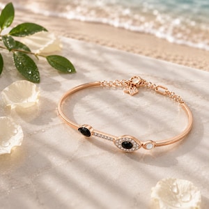 May include: A rose gold-toned bangle bracelet featuring a central eye-shaped motif encrusted with clear crystals and a black stone center. The design includes a smaller black stone, a white stone, and a delicate chain link section with a small charm.